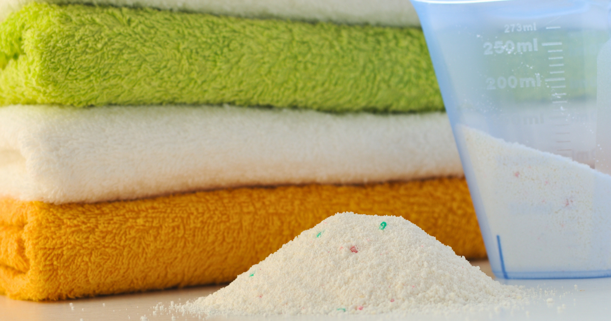 powder vs liquid detergent - The WashRoom Laundry Service Powder laundry detergent and a measuring cup beside folded towels, illustrating powder vs liquid laundry detergent for everyday washing