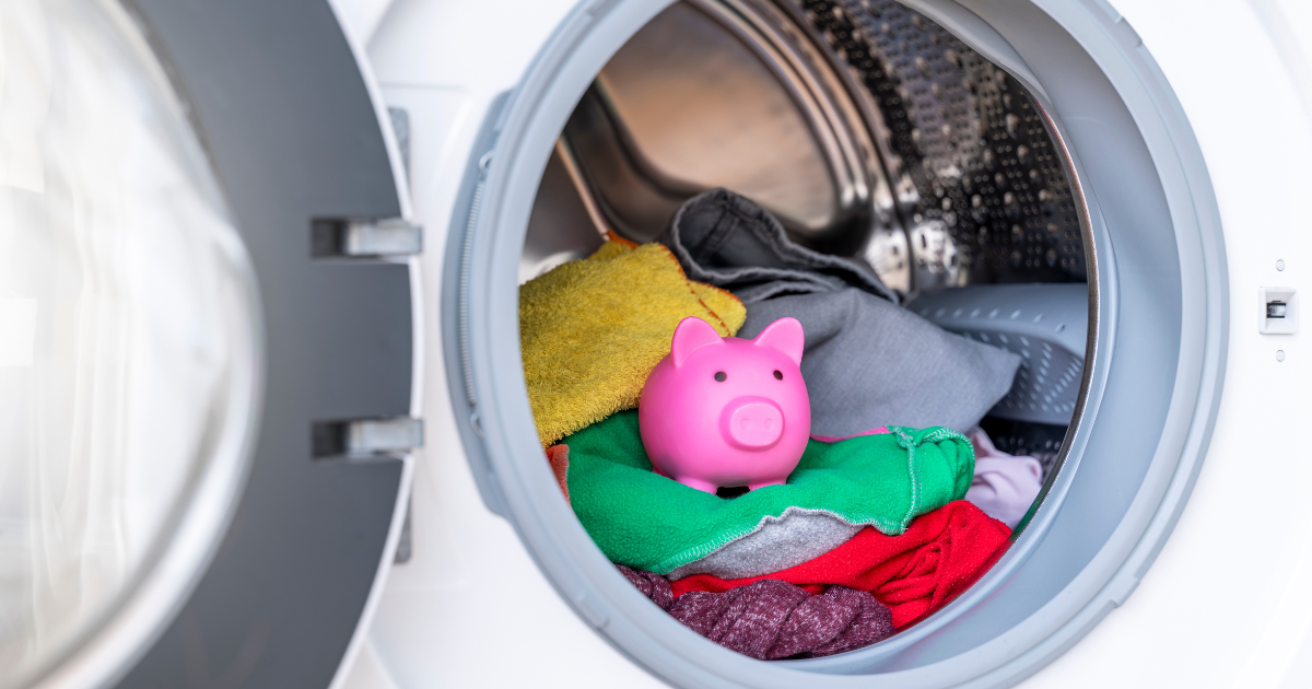 A pink piggy bank sitting on a pile of colorful clothes inside a washing machine, illustrating the hidden laundry cost of doing frequent loads at home.