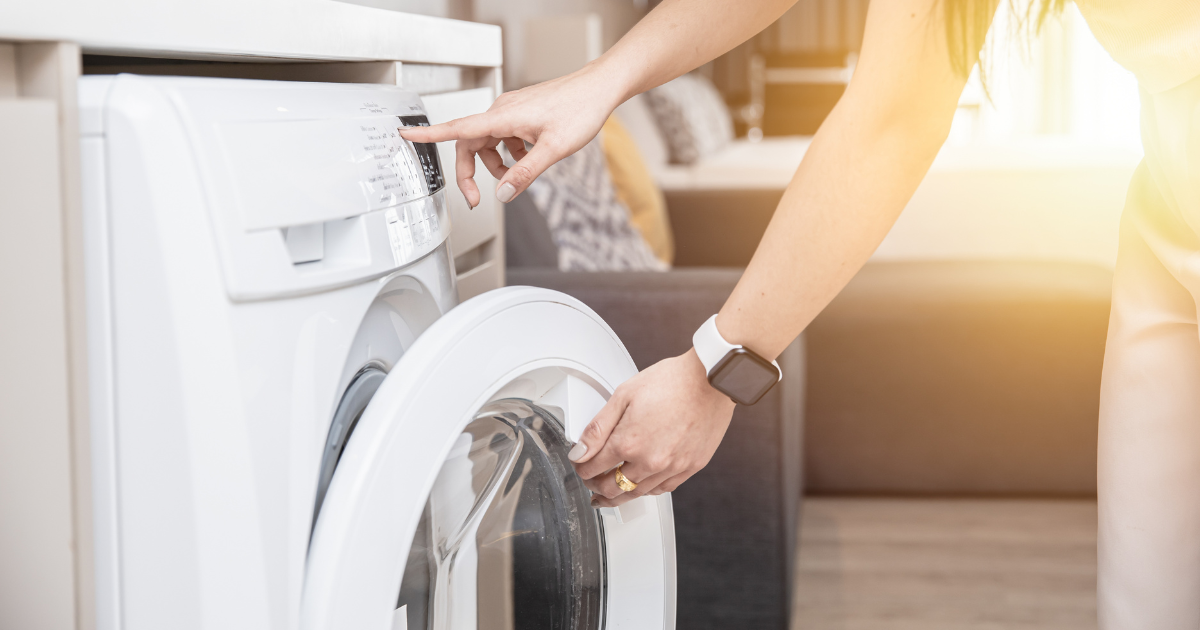 laundry cost - The WashRoom Laundry Service A close-up of a person wearing a smartwatch while starting a washing machine, illustrating the significant time tax involved in a weekly home laundry routine.