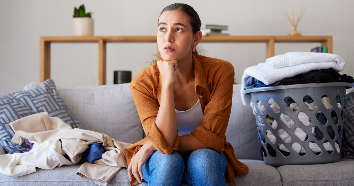 Laundry Help - The WashRoom Laundry Service A frustrated woman sitting on a sofa next to a basket of clothes, looking for laundry help to manage her wash dry fold piles.