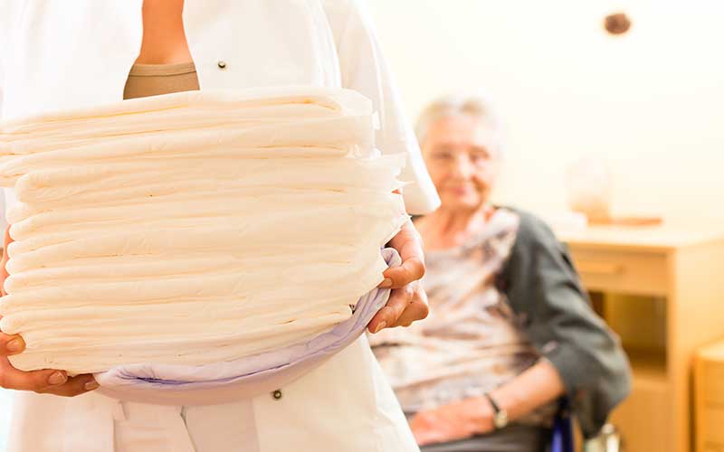 laundry service for seniors - The WashRoom Laundry Service A caregiver holding a stack of clean linens with an elderly woman sitting in the background, showcasing a dedicated laundry service for seniors providing essential senior laundry help.