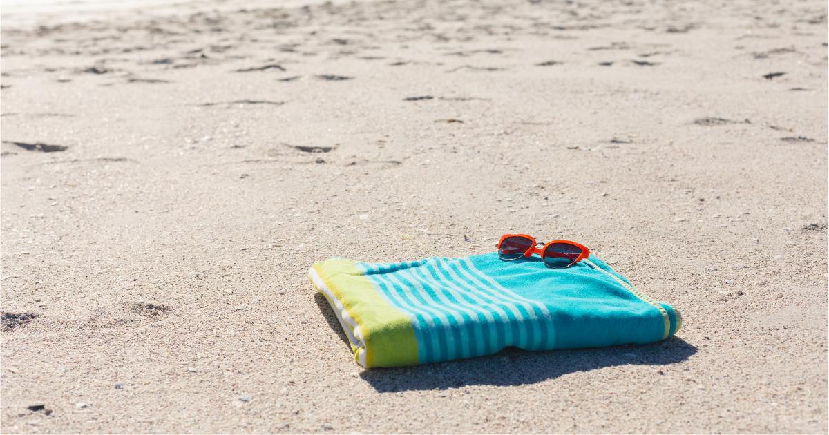 Summer laundry - The WashRoom Laundry Service A bright turquoise and yellow striped beach towel with orange sunglasses on top, lying on light sandy beach, evoking summer laundry needs.