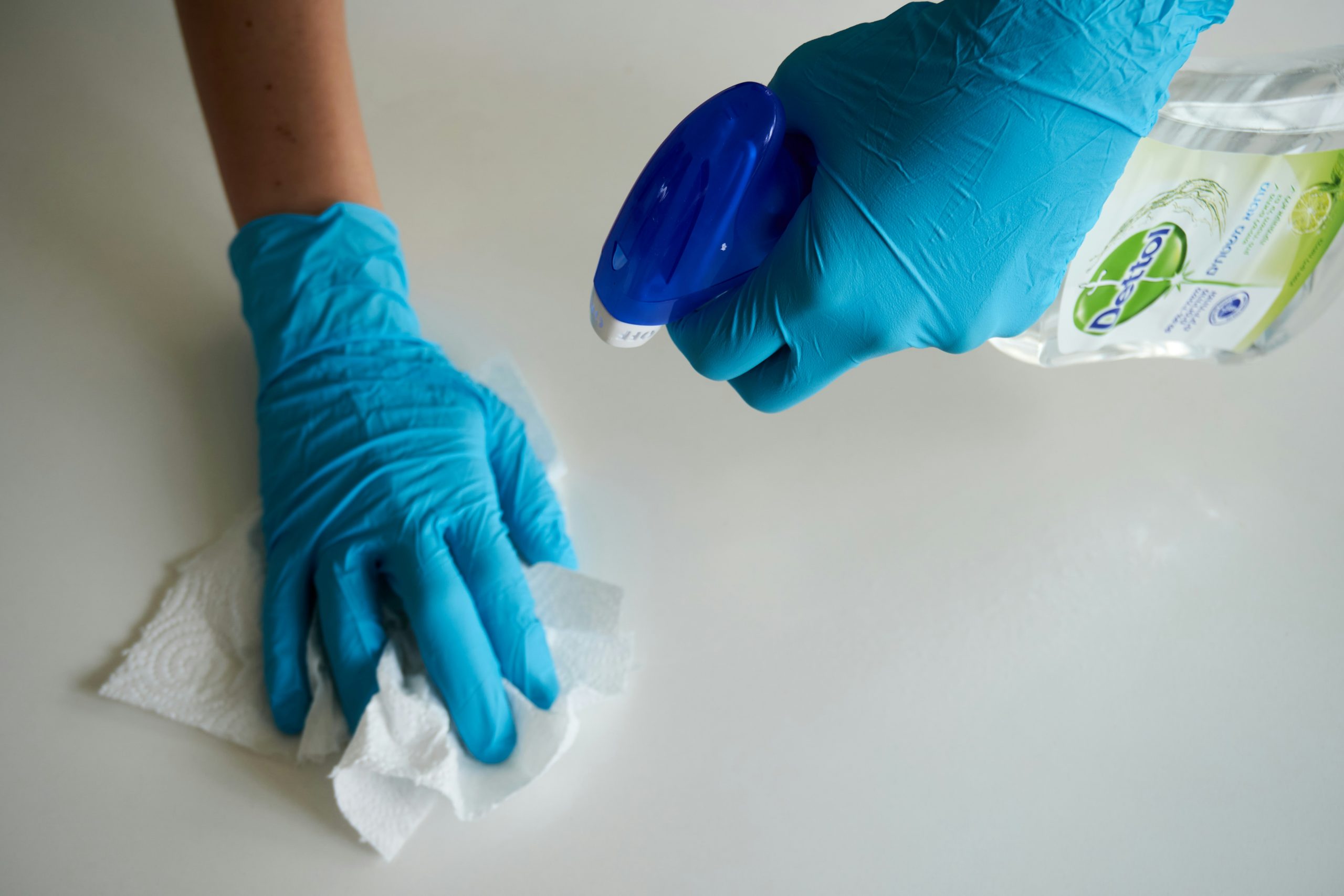 Person wearing blue rubber gloves spraying a white counter with lemon-scented disinfectant cleaner and wiping it with a paper towel, illustrating a step for how to clean your house surfaces.