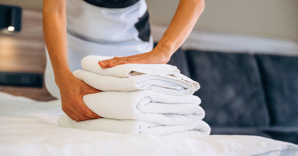commercial laundry - The WashRoom Laundry Service Hotel housekeeper placing a neatly folded stack of fresh white towels on a bed, representing professional commercial laundry service quality