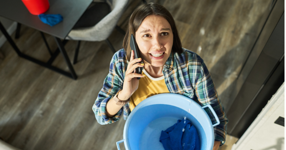 Distressed woman on phone holding a blue bucket to catch a ceiling leak. This image symbolizes the immediate need for professional water damage charlotte cleanup and repair services.