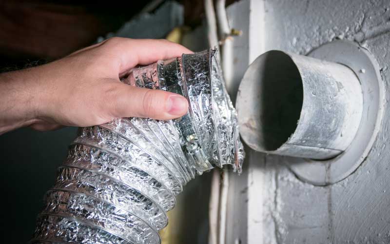 clean a dryer vent duct - The WashRoom Laundry Service A hand disconnecting a flexible silver foil tube to clean a dryer vent duct and remove dangerous lint buildup from the wall connection.